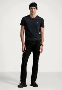 Svart T-skjorte med et subtilt mønster, kombinert med svarte bukser og svarte joggesko. Modellen har på seg en tettsittende svart lue og et armbåndsur.