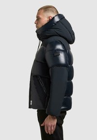 Marine blauwe pufferjack met een glanzende afwerking, oversized capuchon, gewatteerd design en ritssluitingen. Heeft een logo-opdruk op de schouder.
