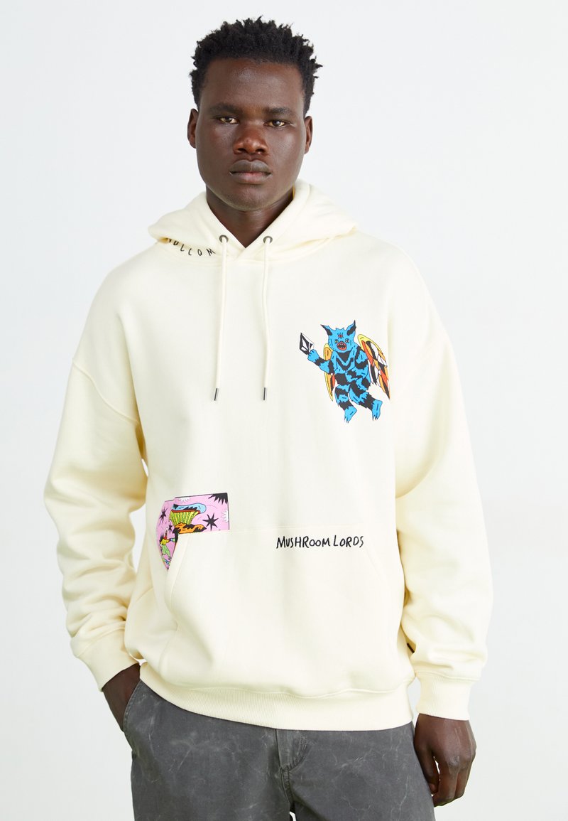 Creme hoodie med grafiske designs: et blåt væsen på forsiden og et pink patch med tropisk billedmateriale. Tekst: "SVAMPEKONGER."