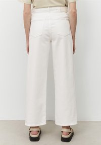 Vita vida byxorna med hög midja och bakfickor, tillverkade av robust denim. I kombination med sandaler med tjock sula, mot en neutral bakgrund.