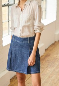 Vaalean beige nappipaita, jossa on kaksi etutaskua, yhdistettynä tummansinisiin farkkuhameeseen, jossa on istuva vyötärö ja hienovaraiset poimut.