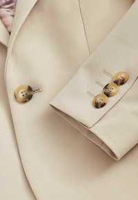 Beige kavaj med slät tyg, en rund knapp på kragen och fyra knappar med marmorering på manschettdetaljen.