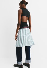 Schwarzes ärmelloses Oberteil mit offener Rückseite, kombiniert mit einer gestreiften Schürze in Grün und Weiß, getragen über dunkelblauen Jeans mit hochgekrempelten Säumen.