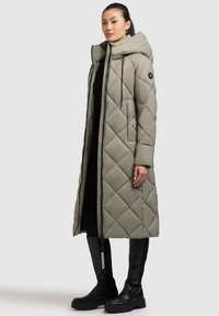 Cappotto invernale lungo, imbottito, grigio, con cappuccio, caratterizzato da un motivo diagonale, zip frontale e tasche laterali. Stivali neri fino al ginocchio completano il look.
