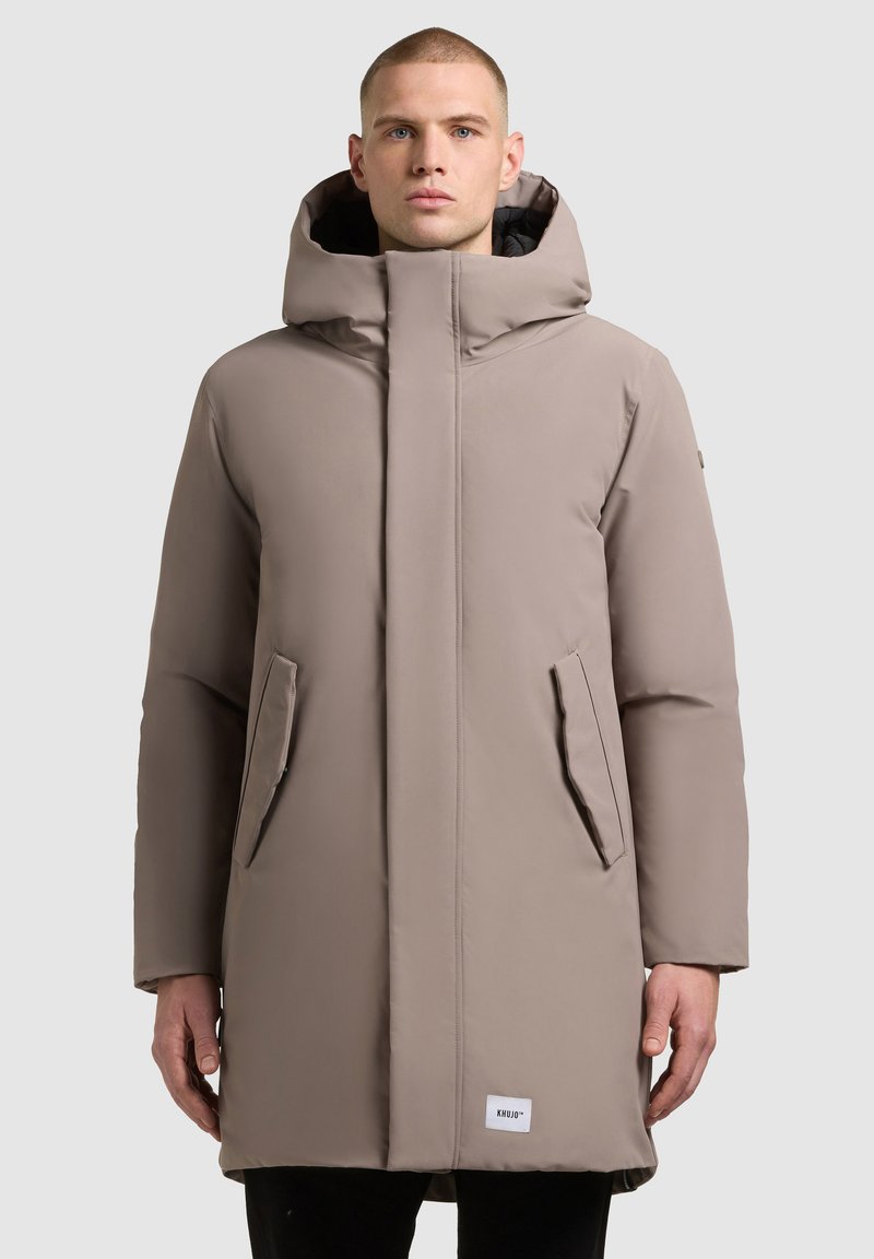 Hosszú, bézs színű steppelt kabát magas gallérral és kapucnival, oldalzsebekkel és kis logócímkével az alján. Simaság és matt felület jellemzi.