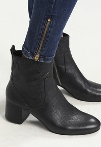 Botas negras hasta el tobillo con un acabado de cuero texturizado, que cuentan con costuras diagonales y un tacón cuadrado. Detalle de cremallera lateral con herrajes dorados.