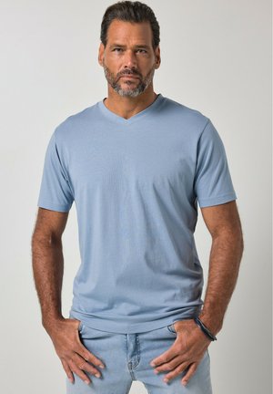 Uomo con la barba che indossa una maglietta azzurra con scollo a V e jeans chiari, in piedi con le mani nelle tasche su sfondo semplice.