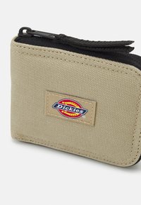 Portafoglio in tessuto marrone con superficie testurizzata, chiusura con zip e un sottolineato patch logo Dickies sulla parte anteriore. Cerniera nera e dettagli in contrasto.