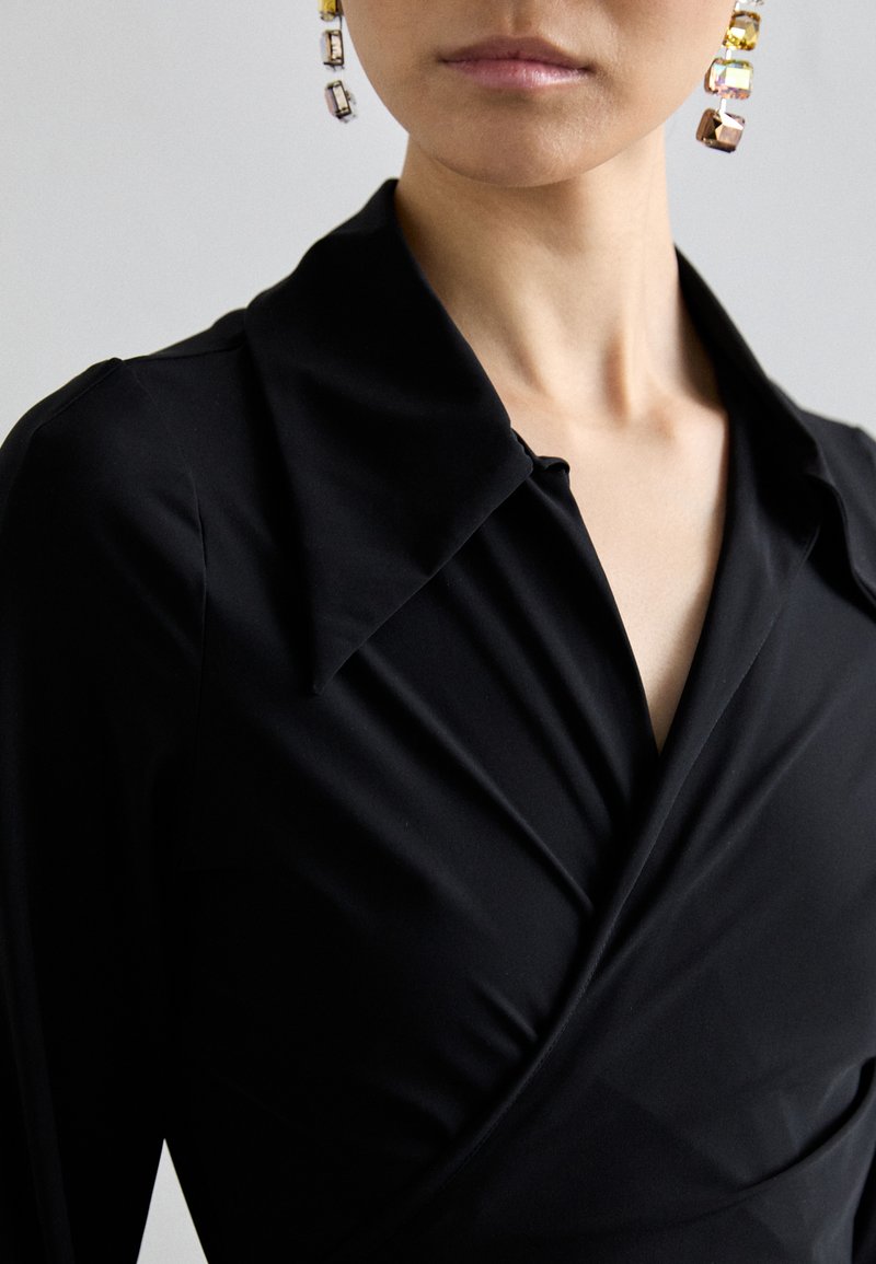 Zwarte wikkeljurk met een opvallende kraag, gemaakt van een gladde, elastische stof. Accentuated met sprankelende oorbellen met meerkleurige stenen.