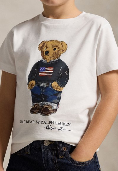 Dziecko noszące białą koszulkę z nadrukiem misia w swetrze z amerykańską flagą i w dżinsach.