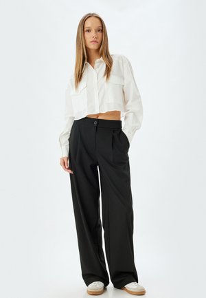 Weißes, kurzes Shirt mit zwei frontalen Taschen, kombiniert mit hochtaillierten schwarzen, weit geschnittenen Hosen und weißen Schuhen mit beigen Sohlen.