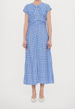 Vrouw draagt een midi-jurk met blauw-wit geruit patroon en met capmouwtjes, gecombineerd met witte pumps met puntige neus, staand tegen een effen achtergrond.