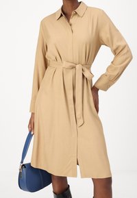 Beżowa sukienka koszulowa z kołnierzykiem, długim rękawem, paskiem w talii i bocznymi rozcięciami; w połączeniu z niebieską torebką i czarnymi botkami.