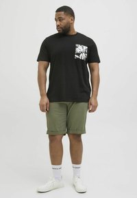 T-shirt noire avec poche à motif, associée à un short vert olive, des baskets blanches et des chaussettes blanches avec un logo. Fond uni.