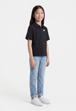 Młoda dziewczyna stojąca, ubrana w czarną koszulkę polo Nike, jasnoniebieskie podwinięte dżinsy i białe trampki, uśmiechająca się na białym tle.