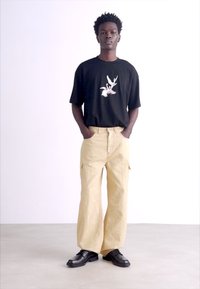 Svart T-shirt med grafiskt tryck, beige cargobyxor med fickor och svarta snörskor. Modellen står självsäkert framför en enkel bakgrund.