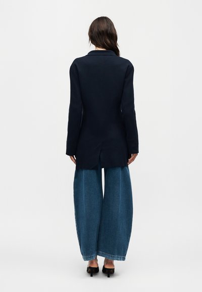 Woman standing facing away, wearing a long black blazer, wide-leg blue jeans, and black high-heeled shoes against a plain background.