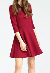 Robe rouge texturée avec manches trois-quarts, corsage ajusté et jupe évasée. Un détail au niveau de l'encolure apporte une touche subtile.