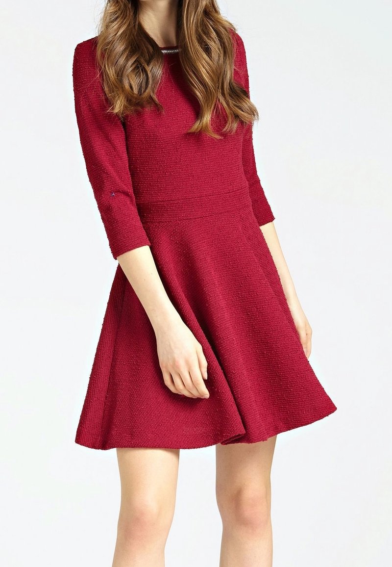Robe rouge texturée avec manches trois-quarts, corsage ajusté et jupe évasée. Un détail au niveau de l'encolure apporte une touche subtile.