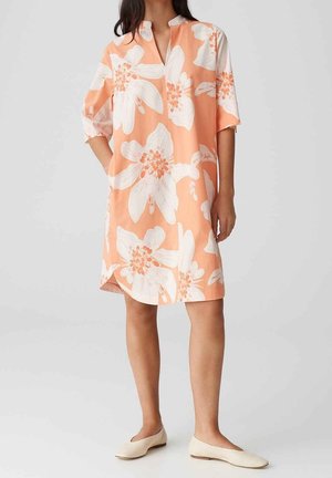 Mujer con vestido color durazno hasta la rodilla con un gran estampado floral blanco, mangas tres cuartos y zapatos planos blancos, de pie frente a un fondo liso.