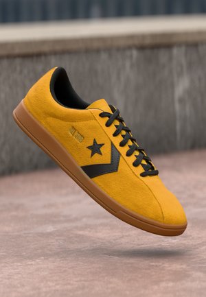 Yellow suede sneaker with black laces and star logo, brown sole, floating above pink concrete ground in urban outdoor setting.