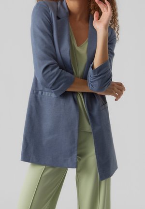 Vrouw die een blauw colbert draagt over een lichtgroene top en broek, met gekruiste armen en een hand dichtbij het gezicht tegen een effen achtergrond.