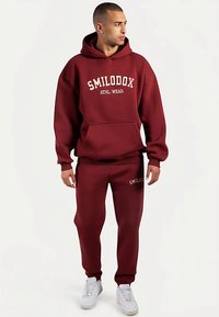 Completo composto da felpa con cappuccio marrone scuro e pantaloni della tuta abbinati, con tasca frontale, scritta "SMILODOX ATHL WEAR" bianca, polsini aderenti e sneakers bianche.