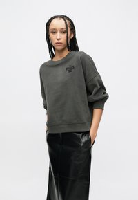 Γκρι φαρδιά φούτερ με στρογγυλή λαιμόκοψη, με ανάγλυφο λογότυπο "The Dance Studio Boss", συνδυασμένο με μαύρο παντελόνι από ψεύτικο δέρμα.