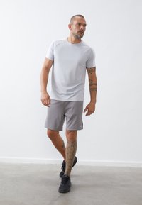 Helles graues Sport-T-Shirt und graue Shorts. Das Shirt hat einen Rundhalsausschnitt und kurze Ärmel. Auffälliges geometrisches Tattoo am Bein. Schwarze Sneakers.