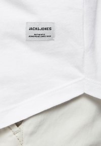 Biela bavlnená tričko s vystúpeným štítkom s nápisom "JACK & JONES - AUTHENTIC JEANSWEAR SINCE 1990" na leme. Viditeľné detaily švov.