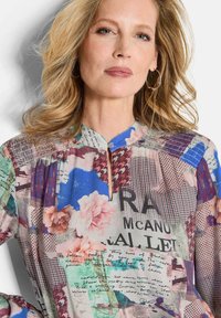 Mehrfarbige Bluse mit floralen und abstrakten Mustern, einem Stehkragen, einem runden Ausschnitt und einem vertikalen Reißverschluss. Leichtes Material.