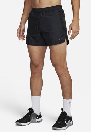 Gros plan sur une personne portant un short de sport noir Nike, des chaussettes blanches Nike montant jusqu'au mollet et des baskets Nike noires et grises, debout devant un fond uni.