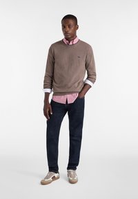 Maglione marrone sopra una camicia colta rosa, jeans scuri e sneakers beige. Le caratteristiche includono un logo sul maglione e le maniche della camicia arrotolate.