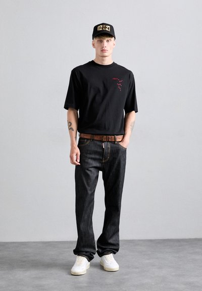 Schwarzes T-Shirt mit kurzen Ärmeln und rotem Text, dunkle Jeans, brauer Ledergürtel, weiße Turnschuhe und schwarze Mütze mit goldener Schrift.