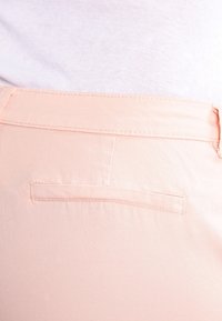 Světlé broskvově zbarvené kalhoty s hladkou texturou. Mají zadní kapsu s obdélníkovou klopou a jemným švem.