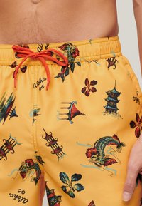 Costume da bagno in tessuto giallo brillante con stampe a tema asiatico colorate, tra cui pagode, pesci e fiori, con un cordoncino rosso.