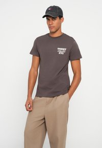 Brązowy bawełniany t-shirt z małym białym logo "TOMMY JEANS NYC" na piersi, w połączeniu z beżowymi luźnymi spodniami i ciemną czapką.