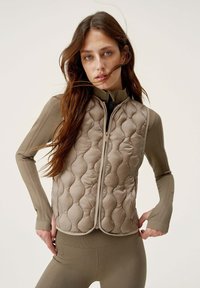 Gilet imbottito senza maniche di color marrone chiaro con un motivo trapuntato. Presenta un colletto alto e una zip frontale. Indossato sopra un top a maniche lunghe coordinato.