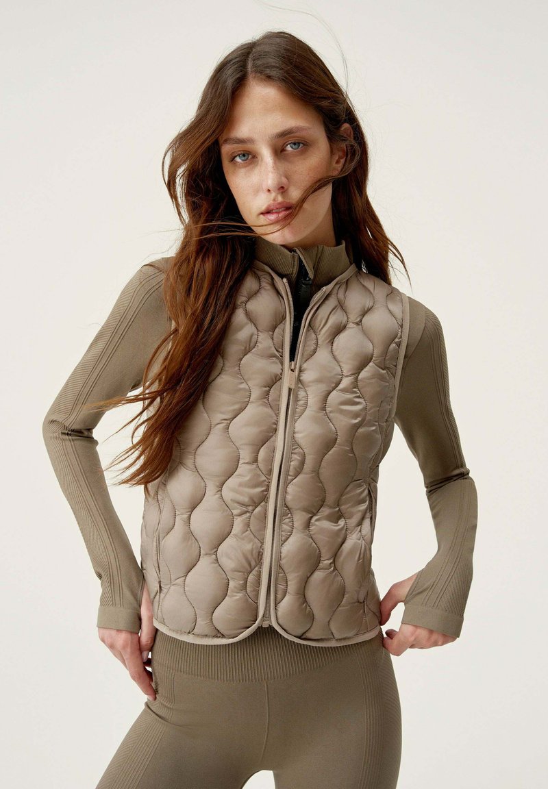 Gilet imbottito senza maniche di color marrone chiaro con un motivo trapuntato. Presenta un colletto alto e una zip frontale. Indossato sopra un top a maniche lunghe coordinato.