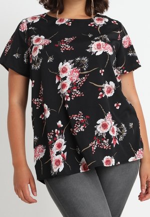 Blouse noire à manches courtes avec imprimé floral rose et blanc portée avec un pantalon gris, montrée du milieu du torse jusqu'aux hanches.