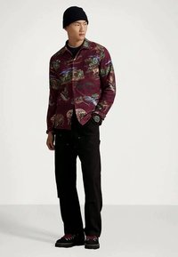 Młody mężczyzna w czarnym beanie, wzorzystej bordowej kurtce, czarnych spodniach i czarnych butach z czerwonymi sznurowadłami, stojący z jedną ręką w kieszeni.