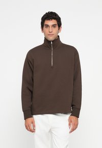 Man som bär en brun långärmad tröja med halvdraget krage och vita byxor, stående mot en enkel vit bakgrund.