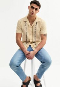 Short-sleeve beige shirt with textured fabric and black floral embroidery down the front, paired with light blue jeans and black sandals.