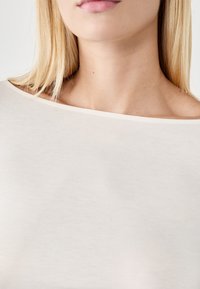 Off-white, dopasowany top z szerokim dekoltem i gładką fakturą. Zbliżenie ukazuje miękki materiał i subtelne odcienie skóry.