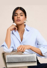 Una donna siede con sicurezza, indossando una camicia azzurro chiaro, mentre tiene una borsa argentata, con uno sfondo neutro che mette in risalto la sua espressione.
