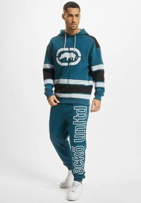 Teal fleece hette med en hvit dyrelogo, svart og lyseblå striper; kombinert med matchende teal joggebukser med stor hvit tekst.