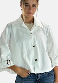 Chaqueta blanca corta en una tela suave con un gran cuello, mangas largas, botones dorados y puños ajustables con un detalle de hebilla negra.