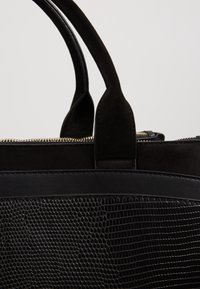 Gros plan d’un sac à main noir avec un corps en cuir texturé, des poignées en daim et une fermeture éclair dorée visible le long du bord supérieur.