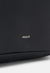 Prodotto in pelle nera con superficie strutturata. Marchio "HUGO" in argento vicino al fondo. Design minimalista con contorno liscio.