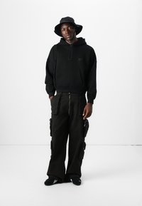 Sort hættetrøje med kengurulomme, sorte cargo-bukser med flere lommer og en sort bucket hat.
